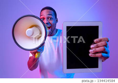Excited African American Guy With Loudspeaker Holding Digital Tablet With Blank Black Screen, Making Announcement While Standing In Vivid Neon Light Over Purple Studio Background, Mockup Image 134414584