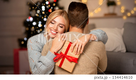Thankful woman holding Christmas gift and hugging her man, celebrating xmas together at home, empty space. Young lady happy to get Christmas present from her boyfriend, embracing him and smiling Thankful woman holding Christmas gift and hugging her man, celebrating xmas together at home, empty space. Young lady happy to get Christmas present from her boyfriend, embracing him and smiling 134414610