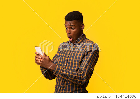 Portraif of shocked funny black man looking at smartphone screen with opened mouth, excited african american guy emotionally reacting to good online offer, standing with isolated on yellow background 134414636