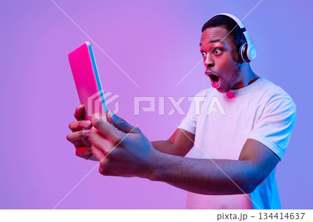 Online Offer. Shocked Black Guy In Wireless Headphones Looking At Digital Tablet With Amazement, Emotional African American Man Standing With Open Mouth Over Purple Background In Neon Light, Panorama Online Offer. Shocked Black Guy In Wireless Headphones Looking At Digital Tablet With Amazement, Emotional African American Man Standing With Open Mouth Over Purple Background In Neon Light, Panorama 134414637