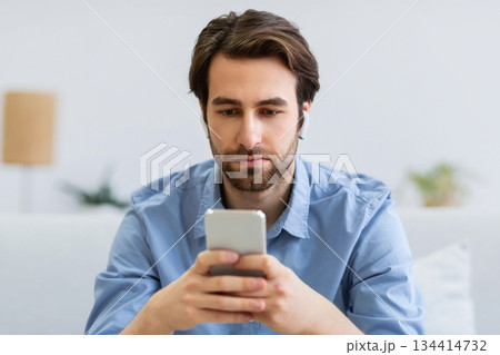 Serious Man Using Smartphone Wearing Earbuds Browsing Internet And Reading News Online Sitting On Sofa At Home. Mobile User Texting On Cellphone In Living Room. Phone Addiction Concept 134414732