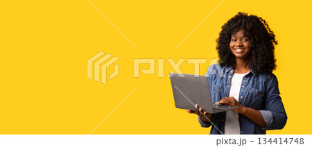 Happy african american young woman holding newest laptop and smiling, yellow studio background, panorama with copy space. Cheerful black lady using notebook, shopping online or websurfing 134414748