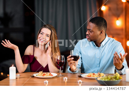 Annoyed African Man Looking At His Girlfriend Talking On Mobile Phone During Romantic Dinner In Restaurant, Complaining About Her Cellphone Addiction. Interracial Couple Having Communication Problems 134414787