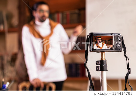 Teacher Blogger Guy Making Video Lesson For Educational Blog Standing Near Blackboard Indoor, Focus On Camera. E-Teaching And Online Learning, Knowledge And Modern Distance Education. Shallow Depth Teacher Blogger Guy Making Video Lesson For Educational Blog Standing Near Blackboard Indoor, Focus On Camera. E-Teaching And Online Learning, Knowledge And Modern Distance Education. Shallow Depth 134414793