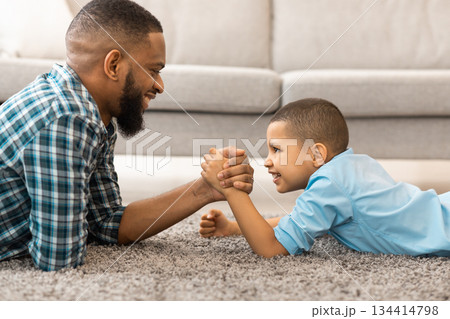 Side View Of Happy Black Father And Son Armwrestling Lying On Floor Having Fun At Home. Little Boy Arm Wrestling Competing With Daddy. Raising A Real Man Concept. 134414798