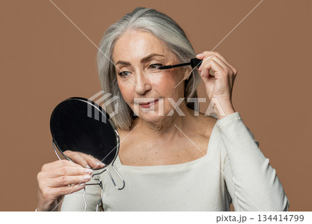 Cheerful senior caucasian female with gray hair applies mascara on eyelashes with brush, looks in mirror, isolated on brown background, studio. Lifestyle, beauty care, natural makeup, daily cosmetics 134414799