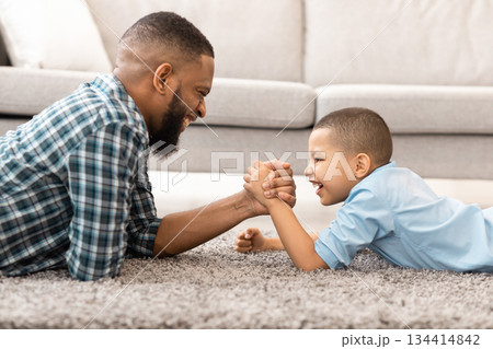Raising A Real Man. Happy African Father And Son Arm Wrestling Competing Lying On Floor And Having Fun At Home. Little Boy Armwrestling With Daddy. Family Bond Concept. Side View 134414842
