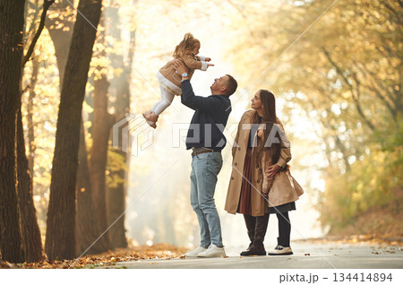 Holding girl in hands. Happy family of mother, father and two daughters are in the autumn park, forest, road Holding girl in hands. Happy family of mother, father and two daughters are in the autumn park, forest, road 134414894