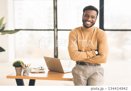Confident Businessman. Happy african guy smiling at camera, sitting on desk with crossed arms. Copy space 134414942