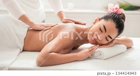 Relaxed asian woman enjoying body massage at spa, getting treatment with professional masseur in beauty parlor, lying on table with closed eyes, having wellness day, side view with free space 134414943