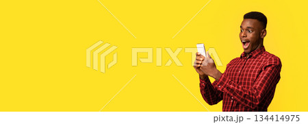 Mobile Offer. Surprised funny black guy looking at smartphone screen with excitement, emotionally reacting to online news, standing with opened mouth isolated over yellow background, copy space 134414975