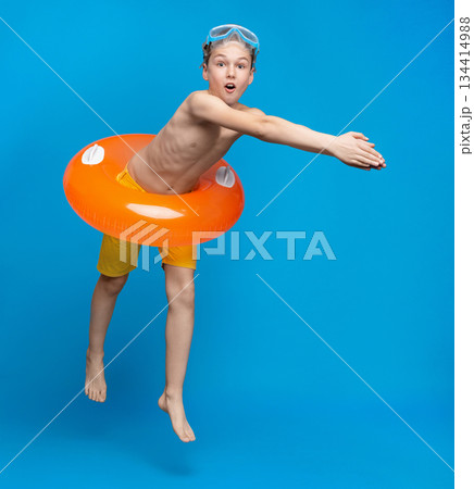 Teen boy in inflatable ring about to dive into swimming pool, blue studio background 134414988