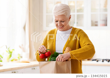Smiling senior lady checking paper bag with grocery ordered from internet during COVID-19 quarantine, kitchen interior, copy space. Happy old woman unpacking purchase. Food delivery concept Smiling senior lady checking paper bag with grocery ordered from internet during COVID-19 quarantine, kitchen interior, copy space. Happy old woman unpacking purchase. Food delivery concept 134415015