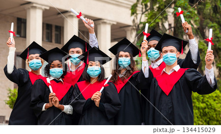 Diverse International Students With Diplomas Celebrating Graduation, happy multiracial group of students in protective face masks and graduation costumes cheerfully posing at university campus 134415029