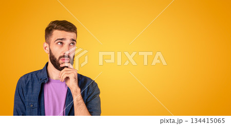 Portrait of pensive young bearded man touching his chin, deep in thought over orange studio background. Doubtful millennial guy thinking and making decision, dreaming about something 134415065