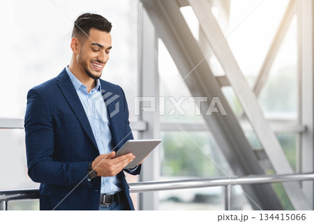 Handsome Young Arab Businessman Using Digital Tablet While Waiting At Airport Terminal, Smiling Middle Eastern Man Working Online Or Browsing Internet On Tab Computer, Enjoying Modern Technologies 134415066