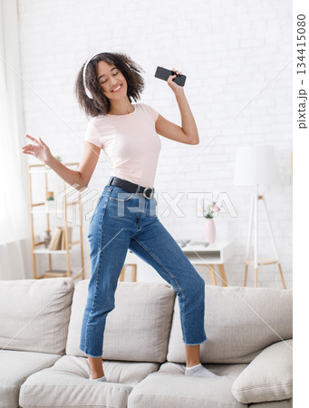 Musical break. African american girl enjoying music and dancing on couch at home during work or study, free space, vertical photo 134415080