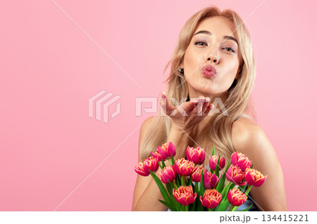Lovely young lady with bouquet of flowers blowing air kiss at camera over pink studio background. Portrait of millennial blonde with bunch of tulips celebrating Woman's Day 134415221