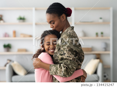 Side view of african american woman in military uniform mother hugging her child happy curly girl teenager at home, mom came back from military service, embracing with her daughter, copy space Side view of african american woman in military uniform mother hugging her child happy curly girl teenager at home, mom came back from military service, embracing with her daughter, copy space 134415247