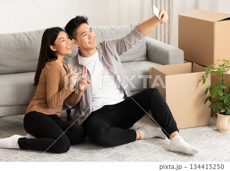 New Home, Technology Concept. Happy asian couple taking selfie with smartphone sitting on floor among cardboard boxes 134415250