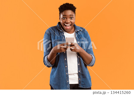 Great Application. Joyful Black Female Using New Smartphone And Mobile App Posing Smiling To Camera Standing Over Yellow Studio Background. Woman Playing Games On Phone Concept 134415260