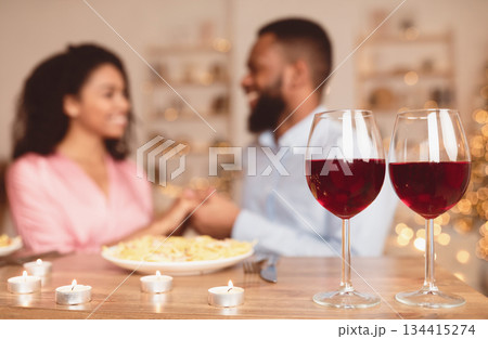 Romantic Date. Selective focus on two glasses of red wine on the table in the foreground. Black couple spending time together at home or cafe, having dinner, holding hands in blurred background Romantic Date. Selective focus on two glasses of red wine on the table in the foreground. Black couple spending time together at home or cafe, having dinner, holding hands in blurred background 134415274