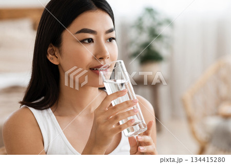 Hydration concept. Young attractive asian woman drinking spring water at home, looking at copy space. Beautiful lady holding glass of mineral water, hydrating her body in the morning 134415280