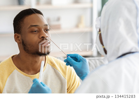 Black guy receiving nasal PCR test while staying home during COVID-19 pandemic, closeup. Unrecognizable doctor in protective suit taking nasal swab for young african american man patient 134415283