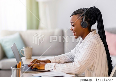 Positive young black woman with headphones sitting in front of laptop, typing on keyboard. female freelancer working from home during COVID-19 pandemic, copy space, side view Positive young black woman with headphones sitting in front of laptop, typing on keyboard. female freelancer working from home during COVID-19 pandemic, copy space, side view 134415287