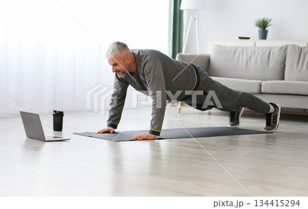 Sporty bearded middle-aged man doing exercises at home, using laptop, watching sport videos online and planking, having fitness class online, copy space. Healthy lifestyle, sport on self-isolation Sporty bearded middle-aged man doing exercises at home, using laptop, watching sport videos online and planking, having fitness class online, copy space. Healthy lifestyle, sport on self-isolation 134415294
