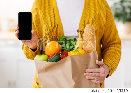 Cropped of woman with shopping bag full of grocery showing smartphone with empty screen, kitchen interior, copy space, mockup. Unrecognizable lady using mobile app for grocery shopping online 134415310