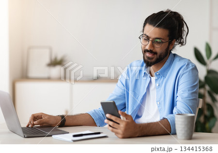 Millennial Arab Freelancer Guy Using Smartphone While Working On Laptop At Desk In Home Office, Browsing New Application Or Messaging With Friends, Enjoying Remote Job Opportunities, Copy Space 134415368