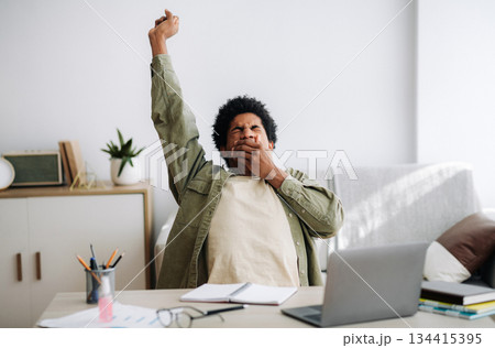 Tired black student yawning and stretching during his remote studies from home. African American youth exhausted from getting ready for test or writing coursework, feeling sleepy in front of laptop Tired black student yawning and stretching during his remote studies from home. African American youth exhausted from getting ready for test or writing coursework, feeling sleepy in front of laptop 134415395