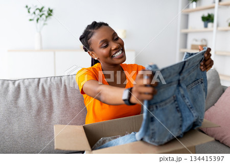 Ordering clothes online. Smiling black female customer opening parcel box, looking at new jeans on sofa at home. Young Afro woman satisfied with internet shopping and delivery service Ordering clothes online. Smiling black female customer opening parcel box, looking at new jeans on sofa at home. Young Afro woman satisfied with internet shopping and delivery service 134415397