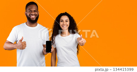 Smiling Multiethnic Couple Showing Cellphone Empty Screen Approving Application Gesturing Thumbs Up Standing Over Yellow Studio Background. We Like This Mobile App. Mockup, Panorama With Copy Space 134415399