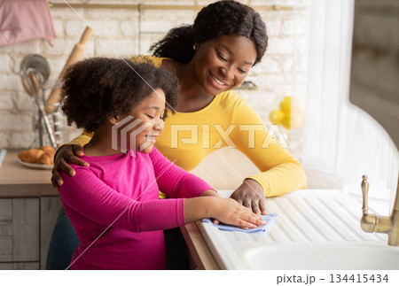 Loving Black Mother Teaching Her Daughter Housekeeping, Cleaning Kitchen Together. Cute Little Girl Wiping Table With Cloth And Smiling, Enjoying Helping With Household Chores, Free Space Loving Black Mother Teaching Her Daughter Housekeeping, Cleaning Kitchen Together. Cute Little Girl Wiping Table With Cloth And Smiling, Enjoying Helping With Household Chores, Free Space 134415434