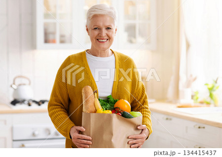 Happy mature woman holding a paper bag full of groceries from the supermarket, kitchen interior. Elderly woman with groceries at home, food delivery during quarantine concept, copy space 134415437