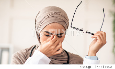Closeup of female doctor woman in hijab removing glasses and rubbing nose bridge, suffering from tired eyes syndrome, clinic interior. Eye fatigue, symptoms and treatments concept 134415508