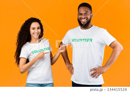 Volunteerting Job. Cheerful Woman Volunteer Pointing Fingers At Male Colleague Standing Over Yellow Background, Smiling To Camera. Diverse Activists Posing Together. Studio Shot Volunteerting Job. Cheerful Woman Volunteer Pointing Fingers At Male Colleague Standing Over Yellow Background, Smiling To Camera. Diverse Activists Posing Together. Studio Shot 134415529