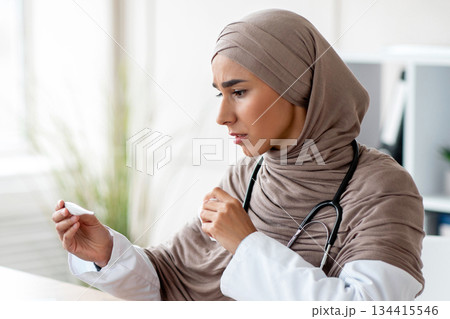 Sick young muslim woman in hijab doctor looking at thermometer, checking fever, sitting at workdesk with laptop in office at clinic, got sick during COVID-19 pandemic, copy space Sick young muslim woman in hijab doctor looking at thermometer, checking fever, sitting at workdesk with laptop in office at clinic, got sick during COVID-19 pandemic, copy space 134415546