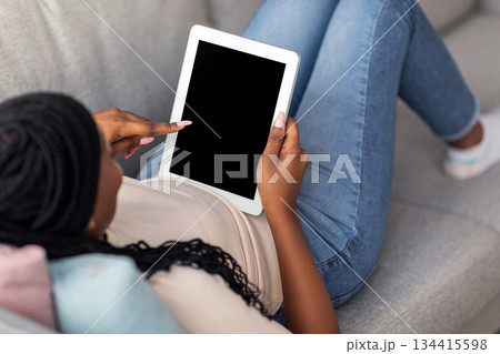Unrecognizable african american woman laying on sofa at home and using modern digital tablet with blank screen, mockup, copy space. Black lady looking at pad with empty display 134415598