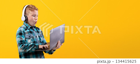 Excited redhead boy with wireless headset playing on laptop, yellow studio background. Amazed kid in casual outfit using modern powerful gadget for video games, playing online, copy space Excited redhead boy with wireless headset playing on laptop, yellow studio background. Amazed kid in casual outfit using modern powerful gadget for video games, playing online, copy space 134415625