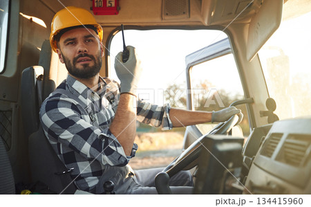 Holding radio, walkie talkie. Construction worker is sitting in the truck cabin Holding radio, walkie talkie. Construction worker is sitting in the truck cabin 134415960