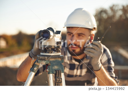 On the radio, with geodetic optical level. Worker is on the construction site at daytime On the radio, with geodetic optical level. Worker is on the construction site at daytime 134415992