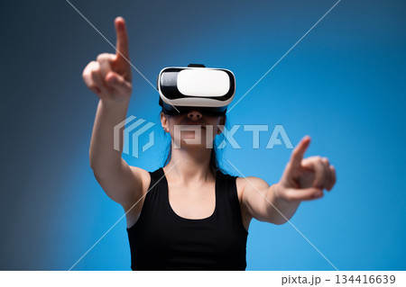 Woman wearing VR headset reaching forward to interact with immersive virtual elements on blue studio background 134416639