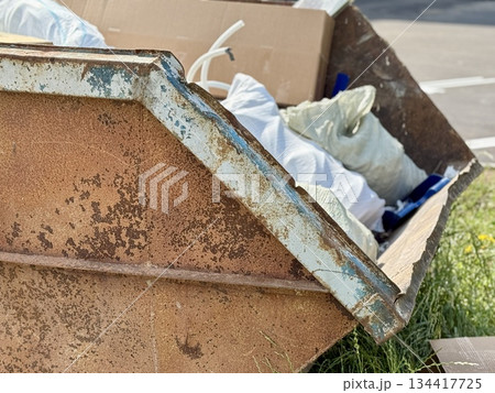 An Old and Rusty Dump Container That is Surrounded by Lush, Overgrown Grass and Weeds. A weathered metal dumpster overflowing with various debris, positioned against a backdrop of lush tall grass. 134417725