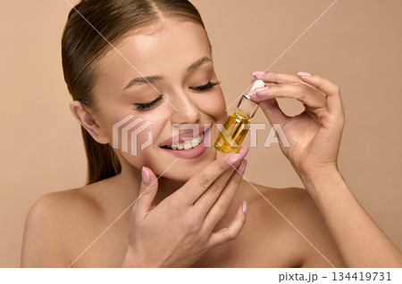 Beauty woman holds serum in studio and smiles while applying skincare for glowing and healthy skin 134419731