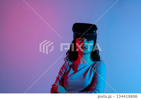 Woman wearing VR headset on forehead looking forward under blended blue and pink neon studio lighting Woman wearing VR headset on forehead looking forward under blended blue and pink neon studio lighting 134419806