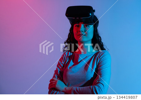 Woman standing with VR headset on her forehead poses confidently under contrasting pink and blue neon lighting Woman standing with VR headset on her forehead poses confidently under contrasting pink and blue neon lighting 134419807