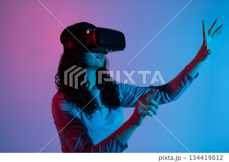 A woman in a VR headset lifts her hand to interact with unseen virtual objects, surrounded by bright neon colors. 134419812